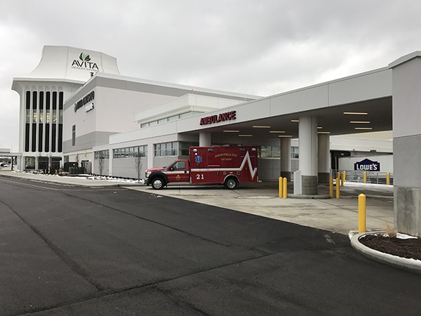 “Covered ambulance entrance at an Avita Health System hospital, with a red emergency ambulance parked under the drive-through canopy labeled ‘Ambulance’ and the main hospital tower visible in the background.