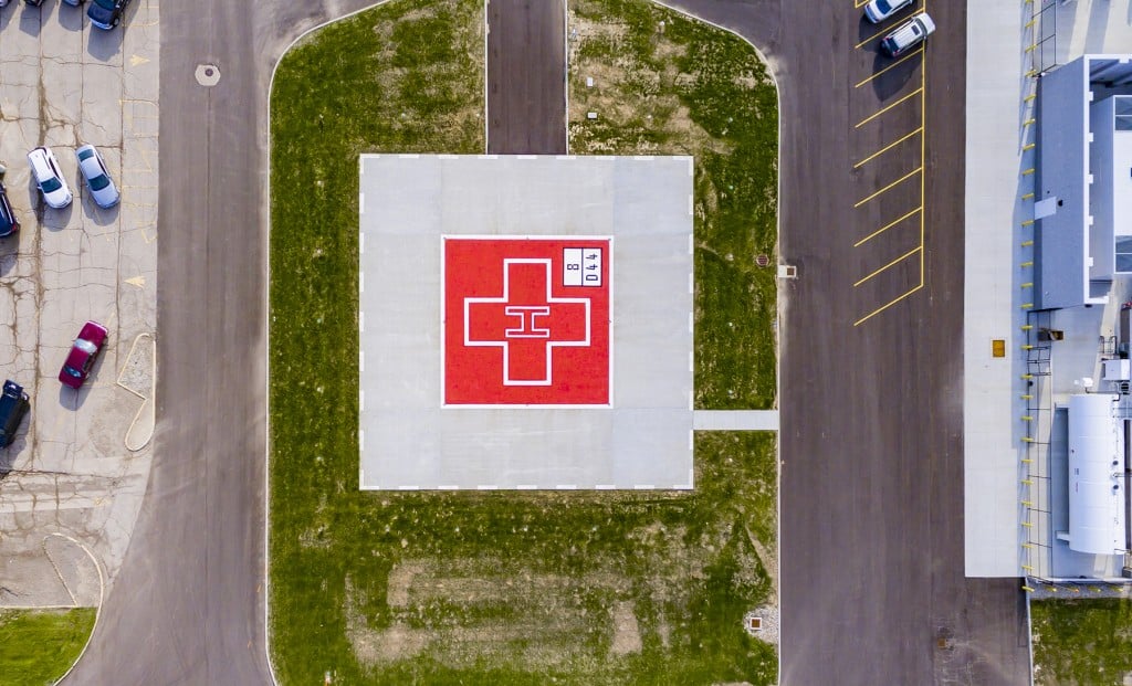 “Aerial view of a rooftop hospital helipad marked with a red square and white cross, surrounded by pavement and adjacent parking areas.