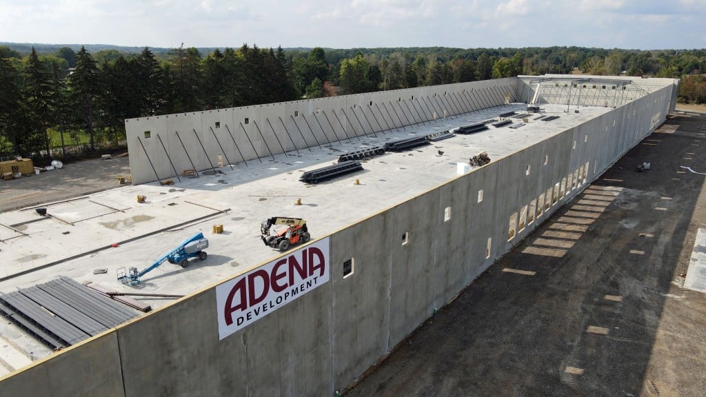 “Aerial view of a long, concrete industrial building under construction with equipment and lifts on the roof and an ‘Adena Development’ sign on the side, surrounded by forested land.