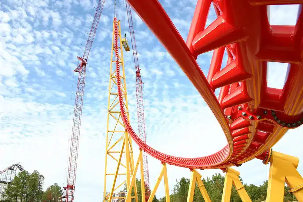 Red roller coaster track diving toward a yellow support tower with cranes in the background.