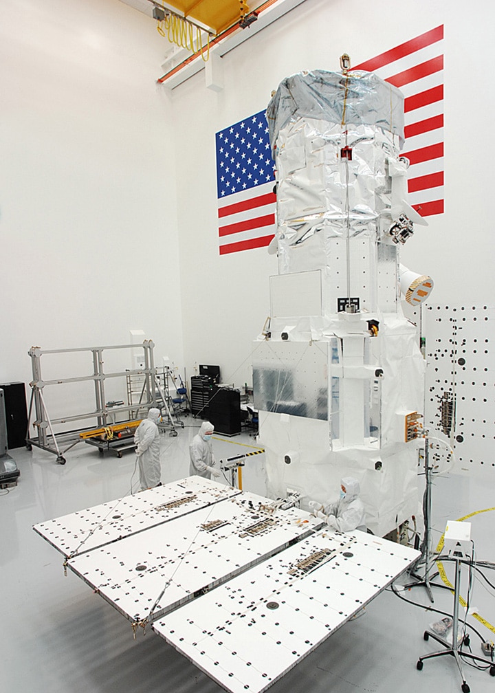 “Technicians in cleanroom suits assembling a large white satellite structure beneath a yellow overhead crane, with an American flag displayed on the wall behind them.