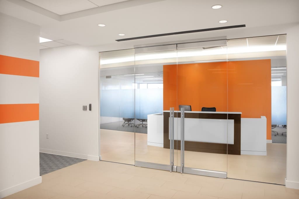 Modern office reception area with a glass sliding door, white front desk, and bright orange accent wall.