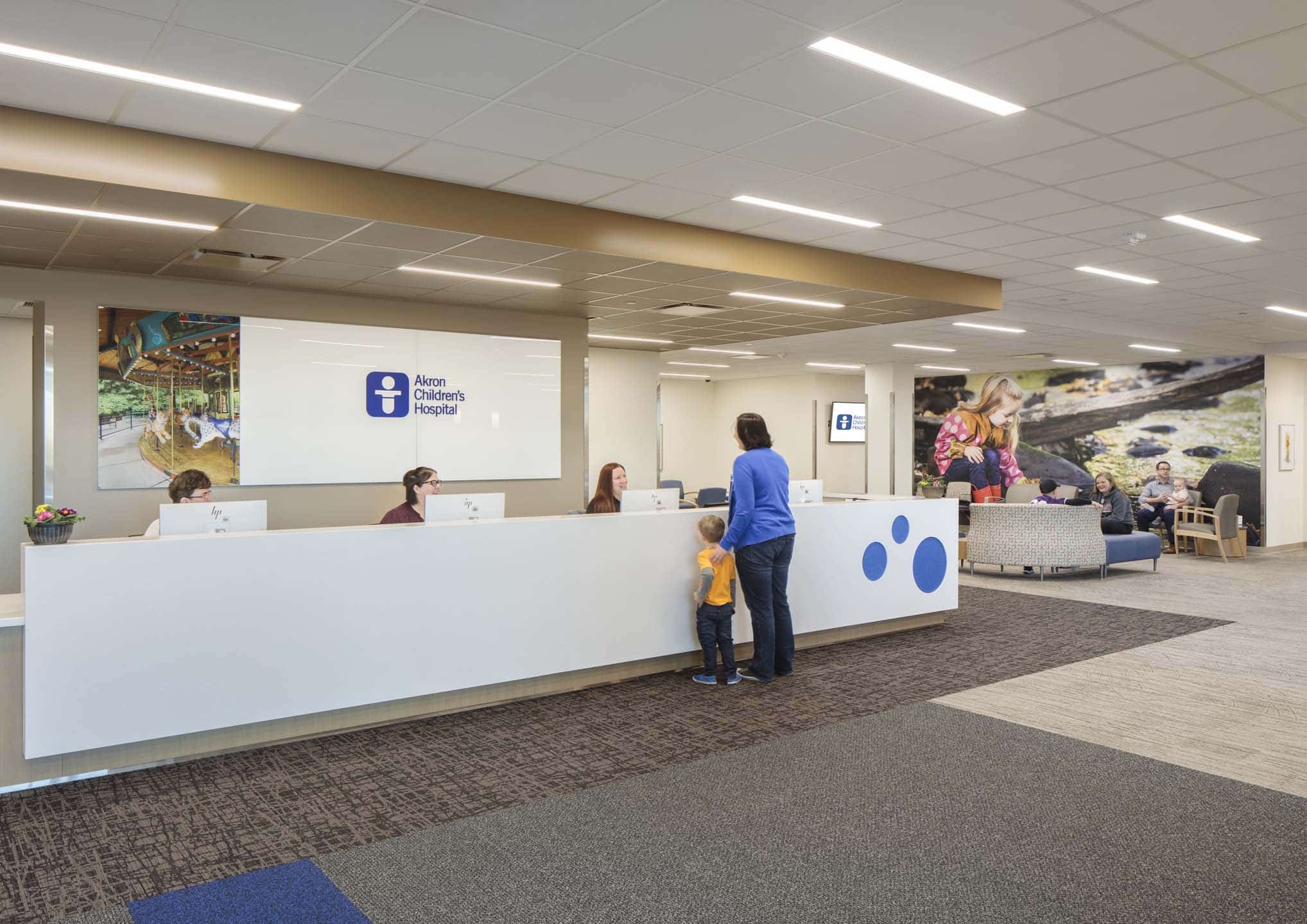 A reception desk in Akron Children’s Hospital with three staff members behind the counter and a child standing with an adult in front of it, with a waiting area and mural in the background.
