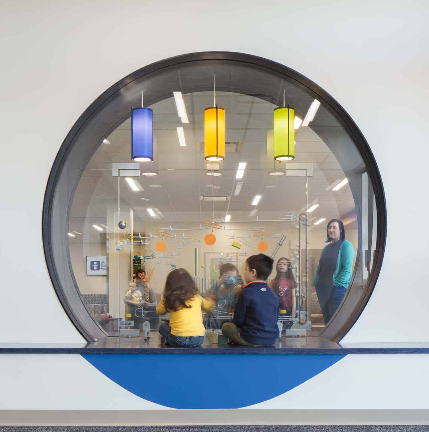 Children sitting on the floor inside a glass-walled room viewed through a large circular window, with an adult standing nearby and three colorful hanging lights above.