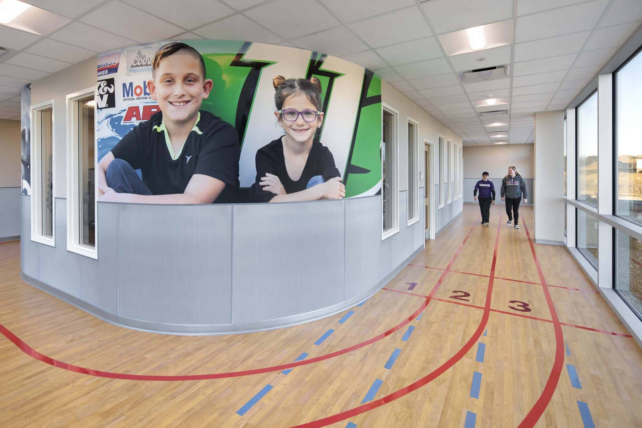 School hallway with a curved half-wall featuring large photos of two smiling children, and two students walking down a floor marked with colorful lines.