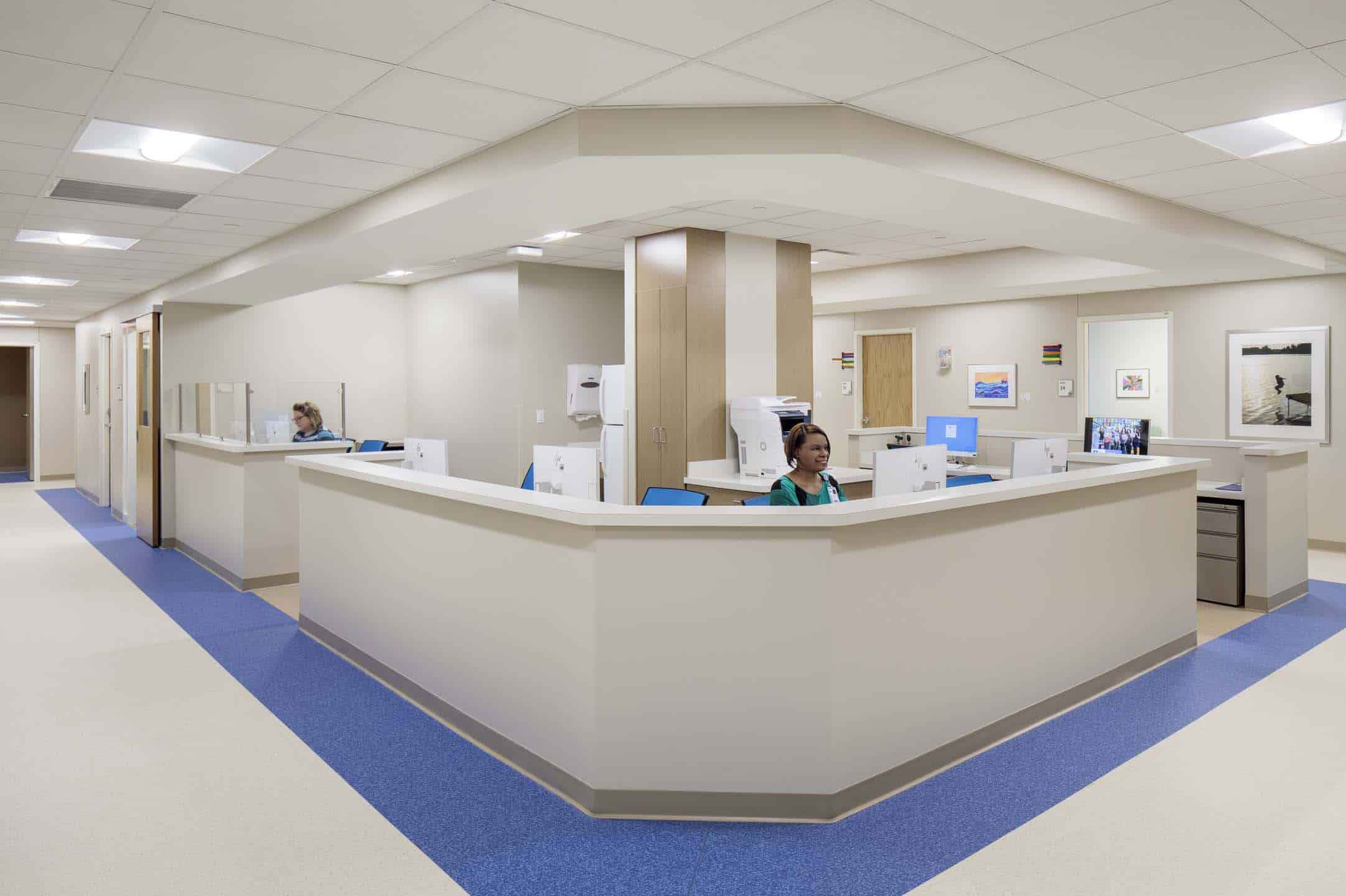 Medical office reception station with a curved counter, two staff members at computers, and a hallway extending to the left.