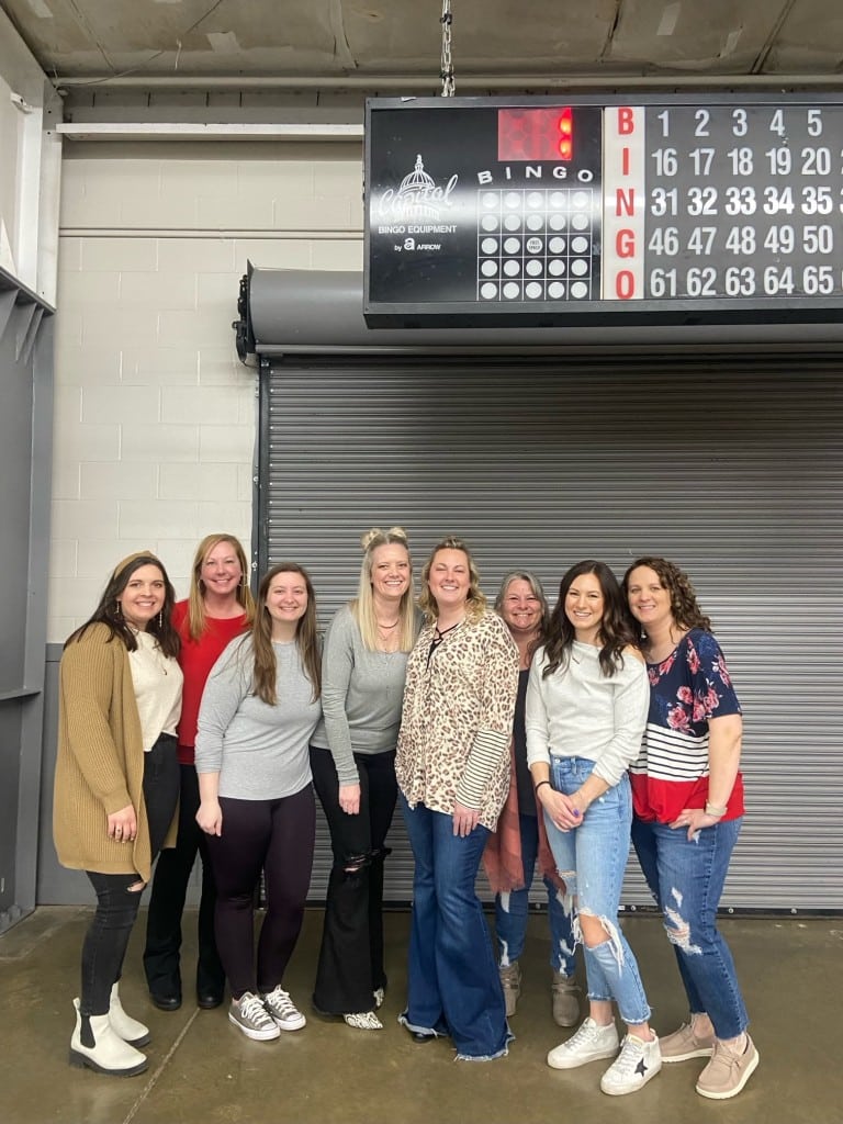 Group of women standing together in front of a closed metal roll-up door beneath a bingo scoreboard.