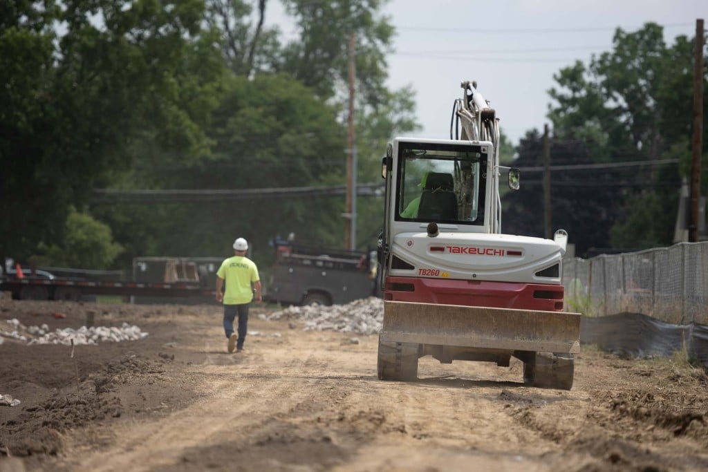 Compact excavator on a dirt construction site with a worker walking ahead.