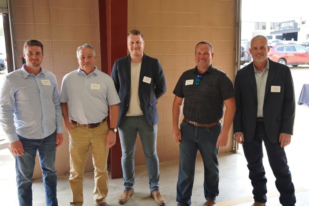Five men standing in a row indoors, smiling at the camera, wearing business-casual attire and name tags in front of a brick wall with an open garage-style door.