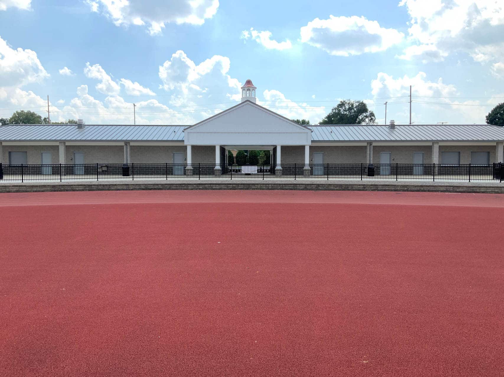 Single-story pavilion building behind a black fence with a red track in the foreground.
