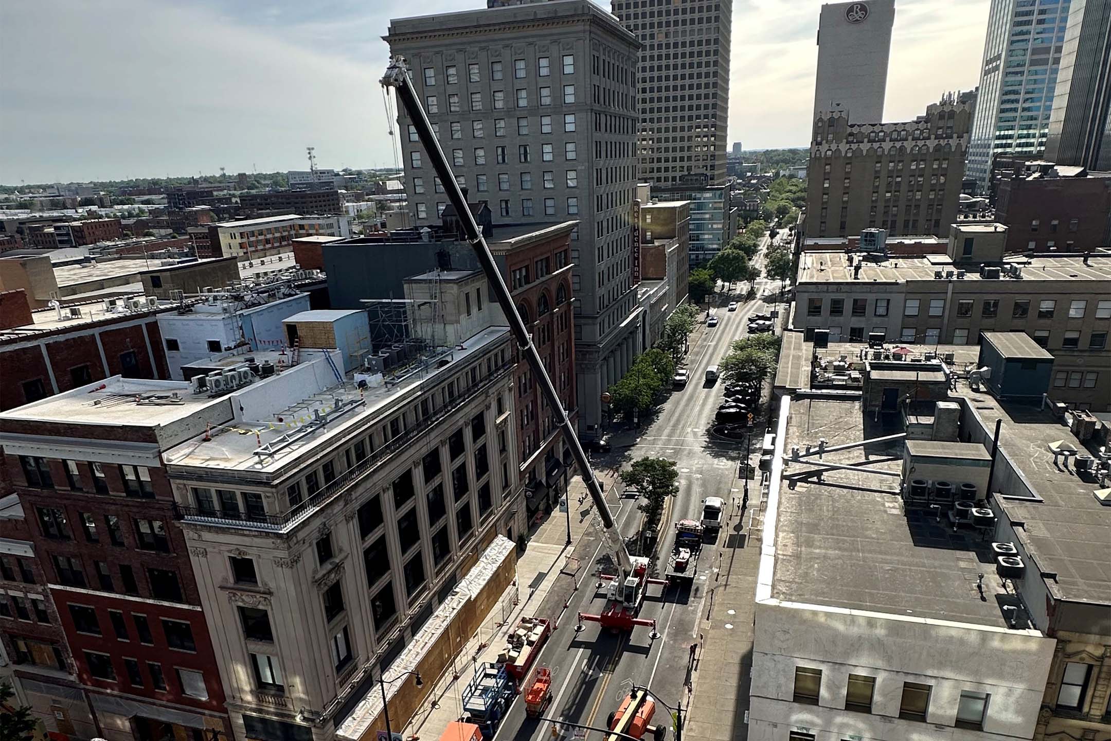 Aerial view of a city street where a long crane boom extends diagonally from a rooftop toward the street below, surrounded by mid-rise downtown buildings and light traffic.