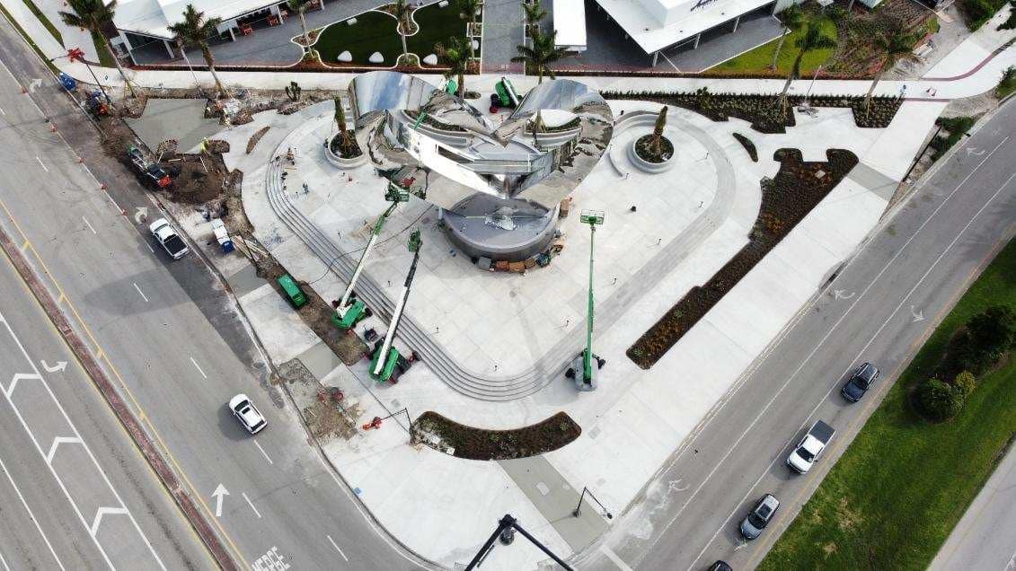 Aerial view of a heart-shaped public plaza under construction, featuring a central sculptural structure, curved seating/steps, and several boom lifts positioned around the site.