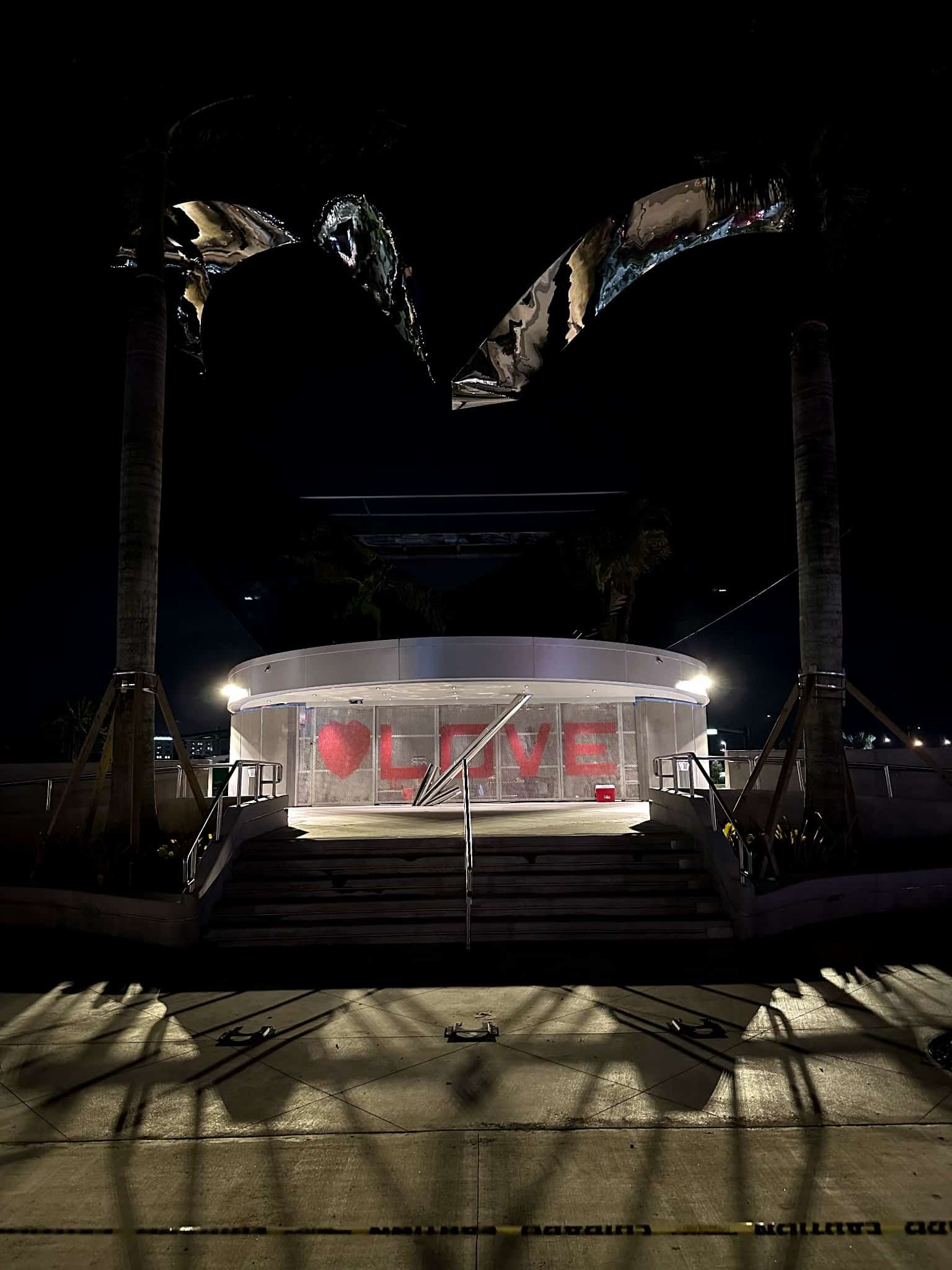 “A circular pavilion at night with illuminated stairs leading up to a glass enclosure displaying a red heart and the word ‘LOVE,’ framed by tall palm trees and curved overhead sculptural elements.