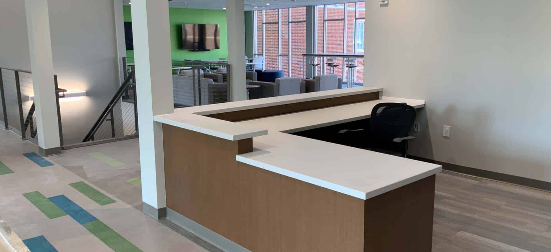 Open reception counter with a modern L-shaped desk overlooking a casual lounge and collaboration area with soft seating, high-top tables, and large windows.