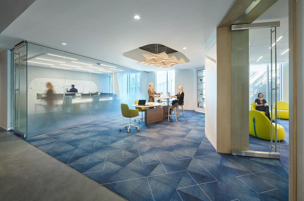 Modern office collaboration space with glass-walled meeting room, high-top table where two people are working together, and a small lounge area with bright green chairs.