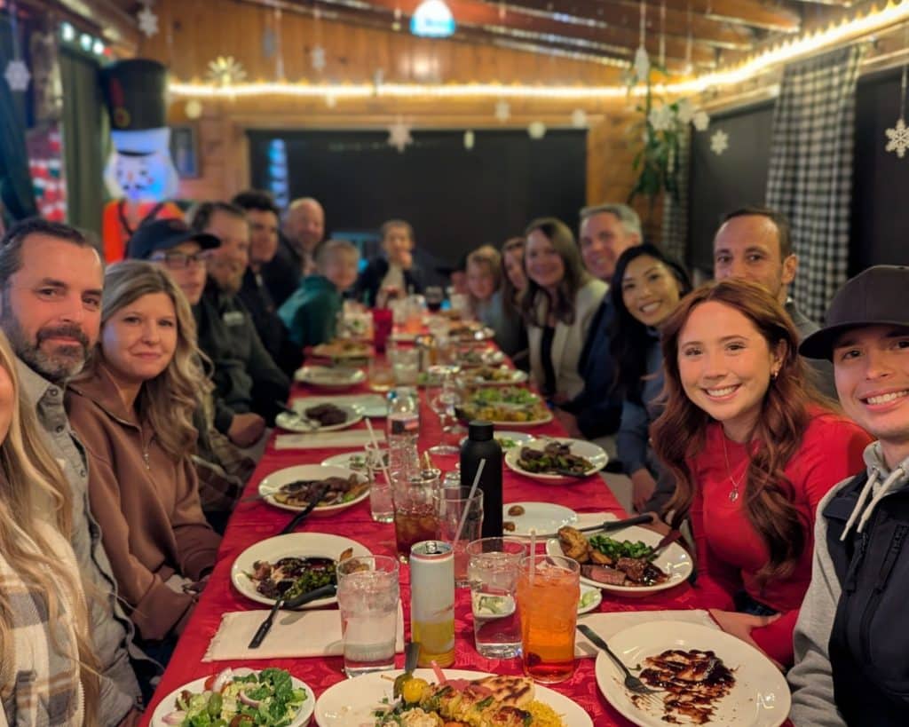 Group of people seated around a long table during a shared meal in a warmly lit, wood-paneled space.