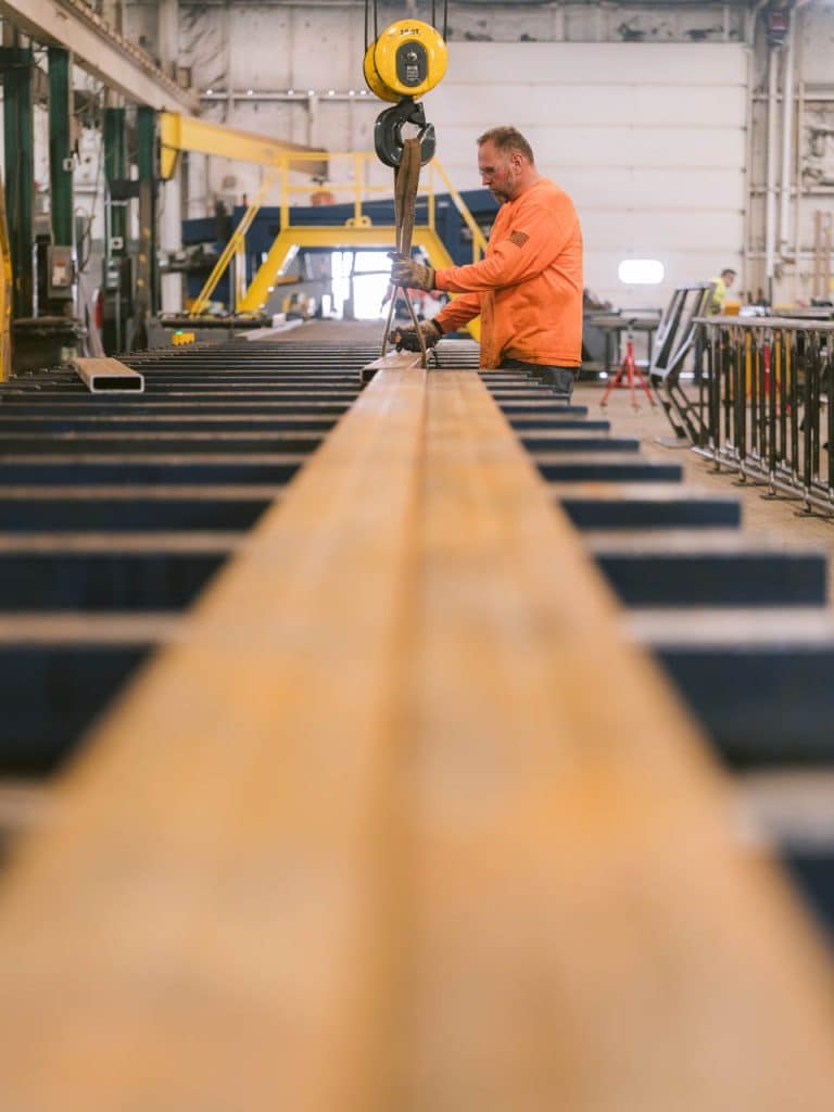 A worker in an orange shirt operating a yellow overhead hoist to lift a long steel beam inside an industrial fabrication shop.