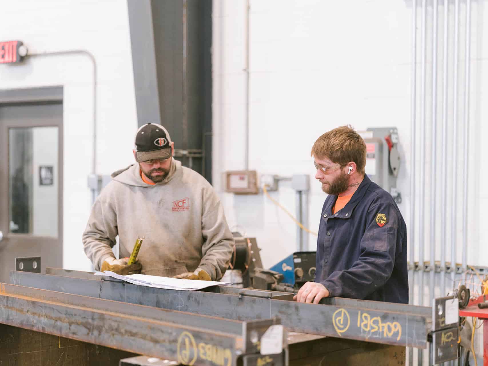 Two workers in safety glasses reviewing plans beside a large steel beam inside an industrial fabrication shop.
