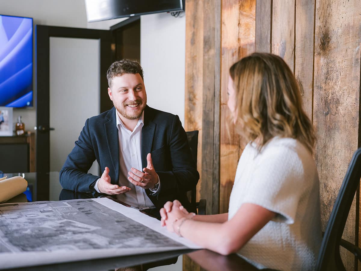 Two people in a professional meeting across a table with building plans.