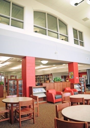 Interior of a school library with round wooden tables and chairs, red accent columns, bookshelves, and soft seating in the background.
