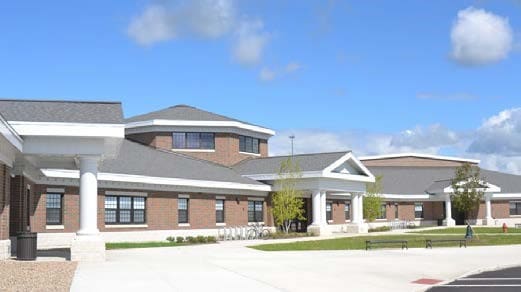 Exterior view of a single-story brick school building with columned entrances and a paved drop-off area in the foreground.
