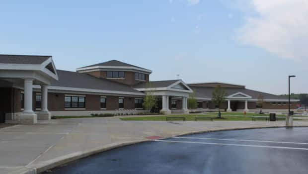 Exterior view of a single-story brick school building with columned entrances and a paved drop-off area in the foreground.