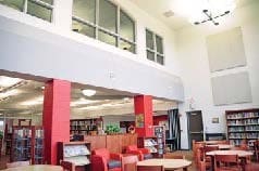 Interior of a school library with round wooden tables and chairs, red accent columns, bookshelves, and soft seating in the background.