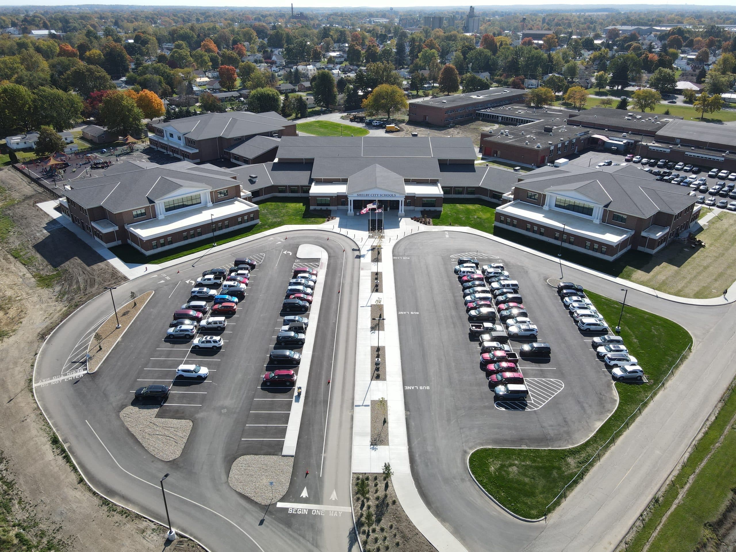 Aerial view of a large school campus with a central entrance, two curved parking lots filled with cars, and surrounding neighborhoods and trees in the background.