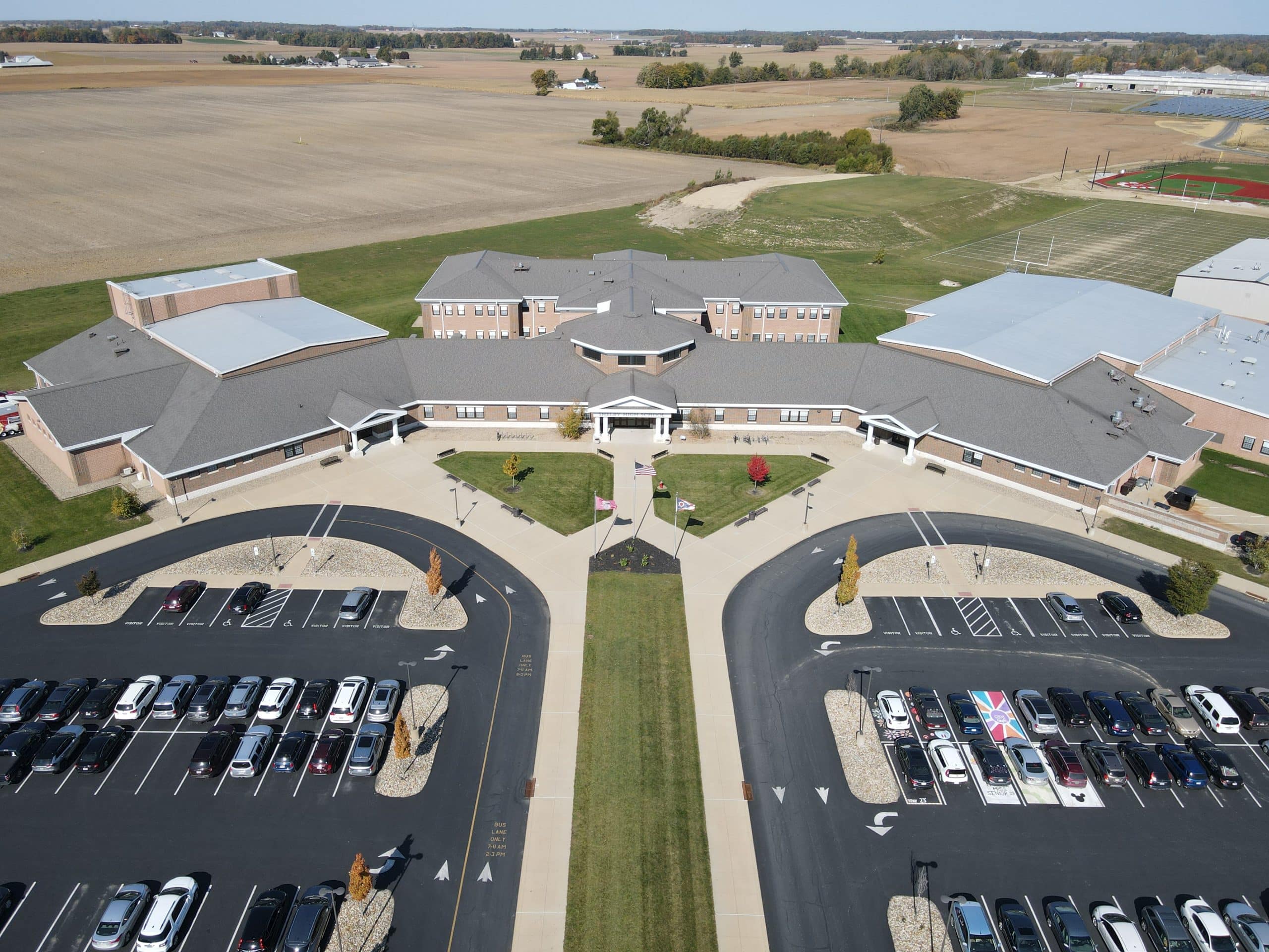 Aerial view of a large single-story school campus with a central entrance courtyard, surrounding parking lots, and open farmland in the background.